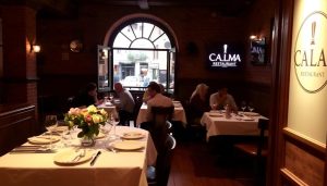 A cozy, dimly lit Spanish restaurant interior, with wooden floors, exposed brick walls, and warm lighting. In the foreground, a table is set with a crisp white tablecloth, classic Spanish tableware, and a centerpiece of fresh flowers. In the middle ground, a group of diners enjoy their meals, engaged in lively conversation. The background features a large, arched window overlooking a picturesque street, with the CALMA restaurant logo visible. The overall atmosphere is one of relaxation and culinary delight.